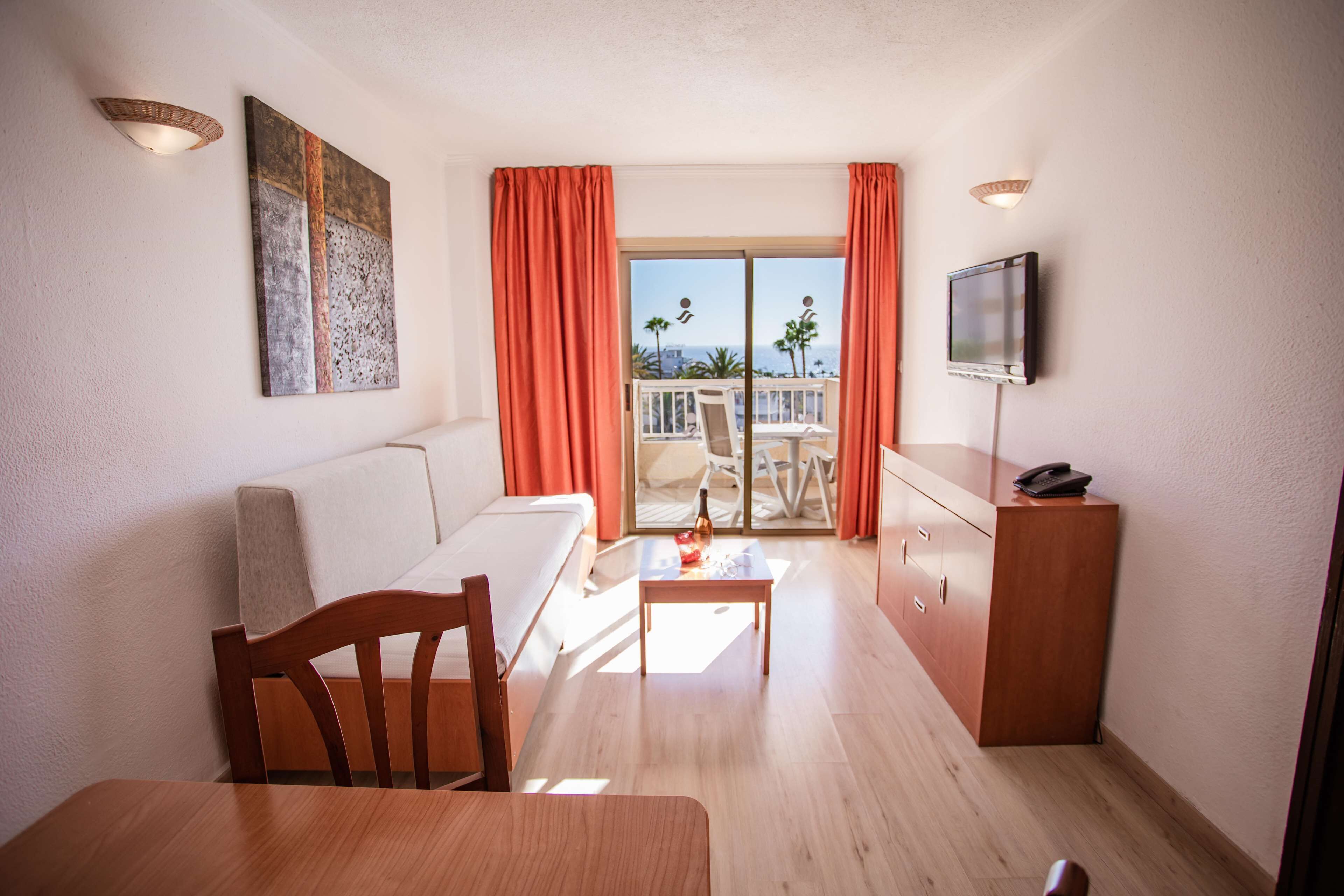 Una sala de estar moderna con un sofá, una mesa de comedor y un televisor. Hay un balcón con vista a palmeras y al océano.