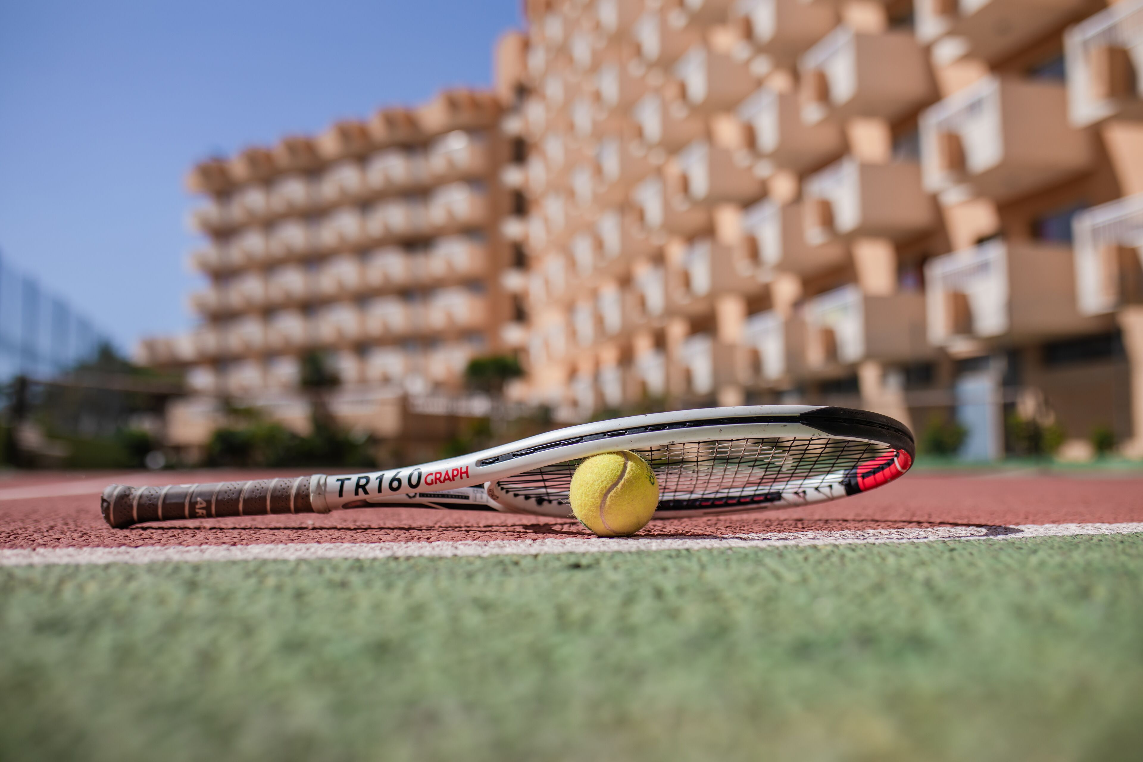 Une raquette de tennis et une balle sur une piste, avec un immeuble à plusieurs étages en arrière-plan.