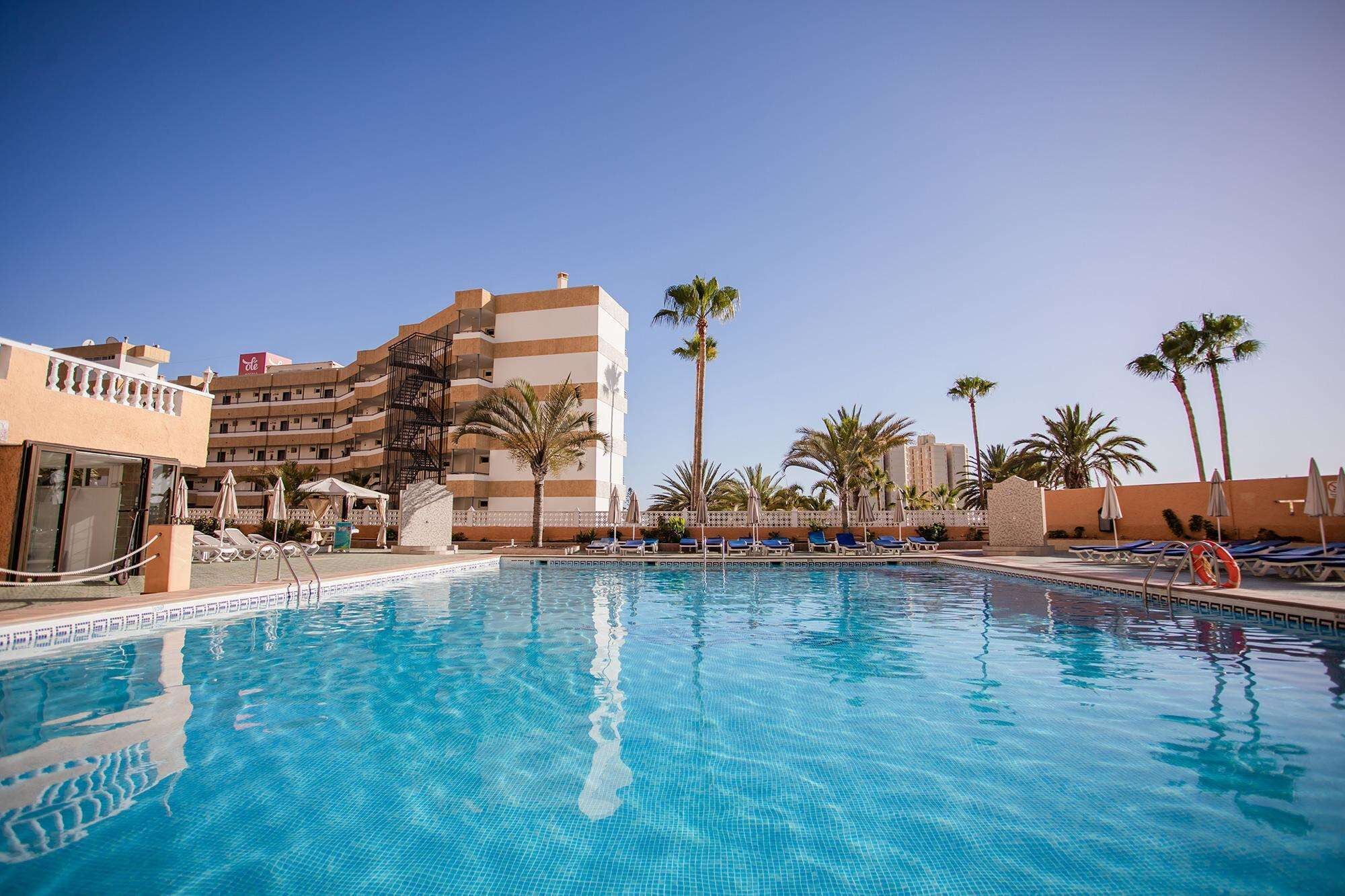 Zona de piscina de hotel con una piscina azul cristalina, sillas de descanso y palmeras.