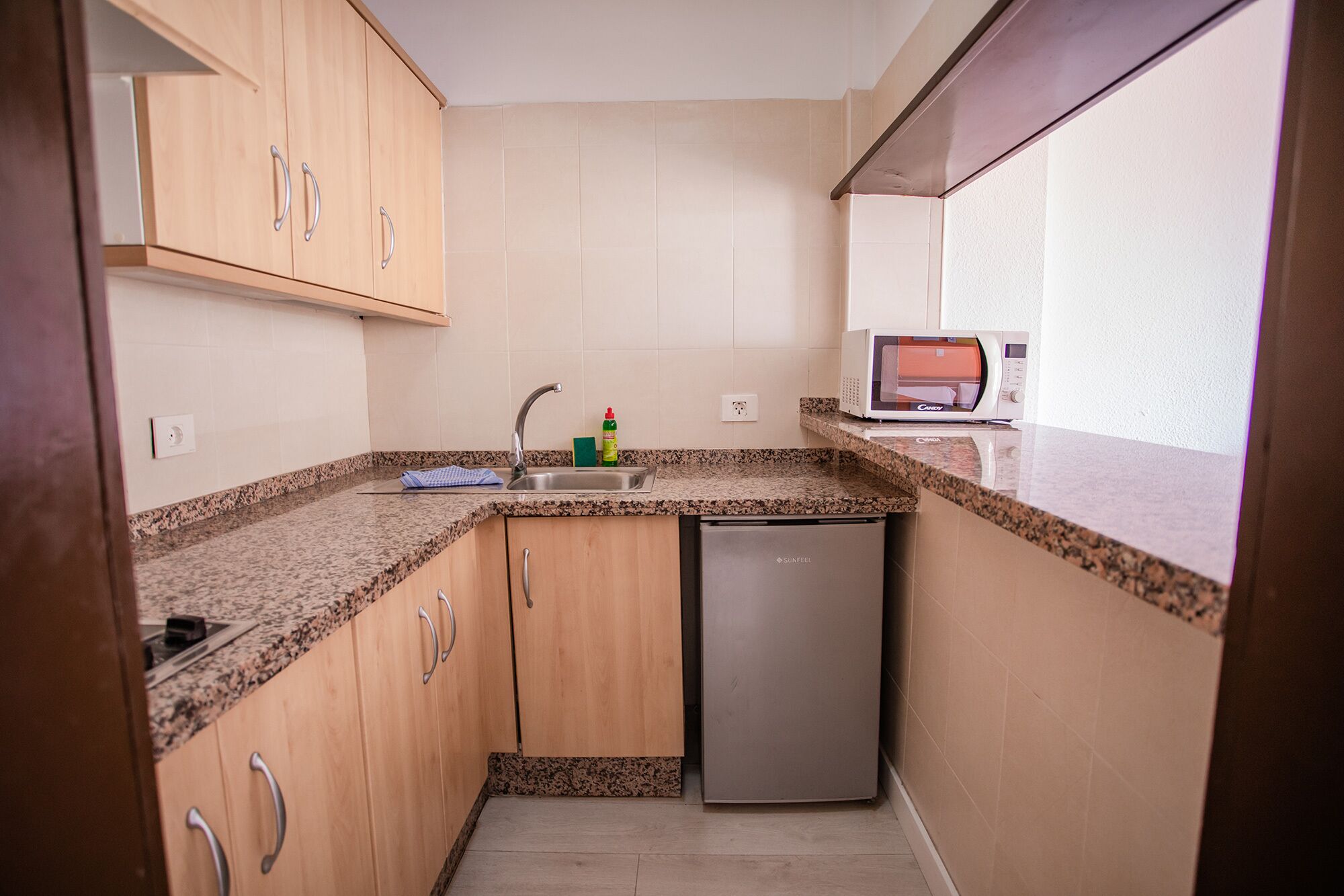 Cocina compacta con armarios de madera, encimera de granito, lavavajillas y microondas.