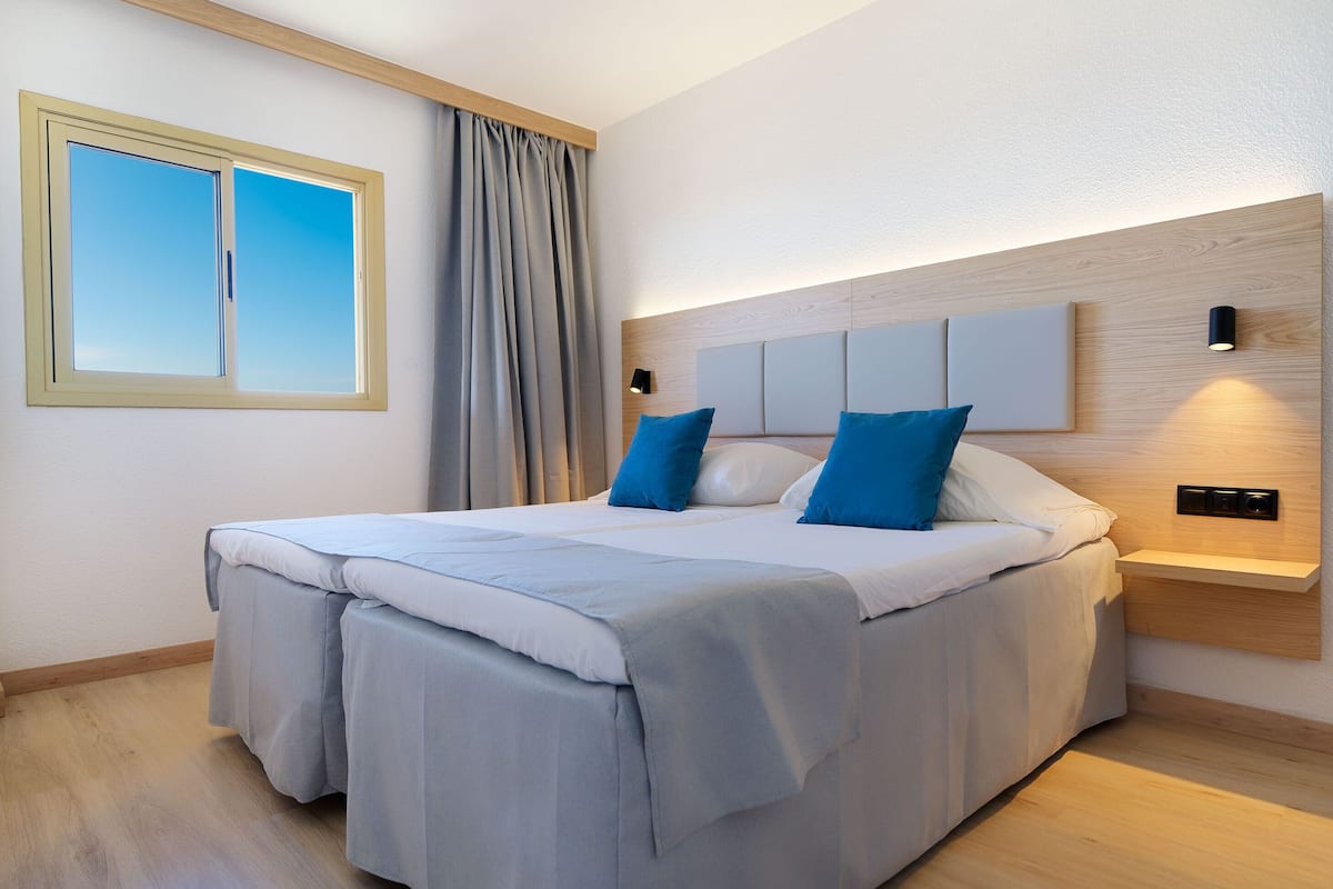 A modern bedroom with a large bed, a headboard with built-in lighting, a window with curtains, and a wooden floor.