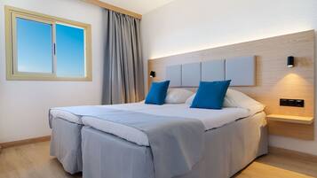 A modern bedroom with a large bed, a headboard with built-in lighting, a window with curtains, and a wooden floor.