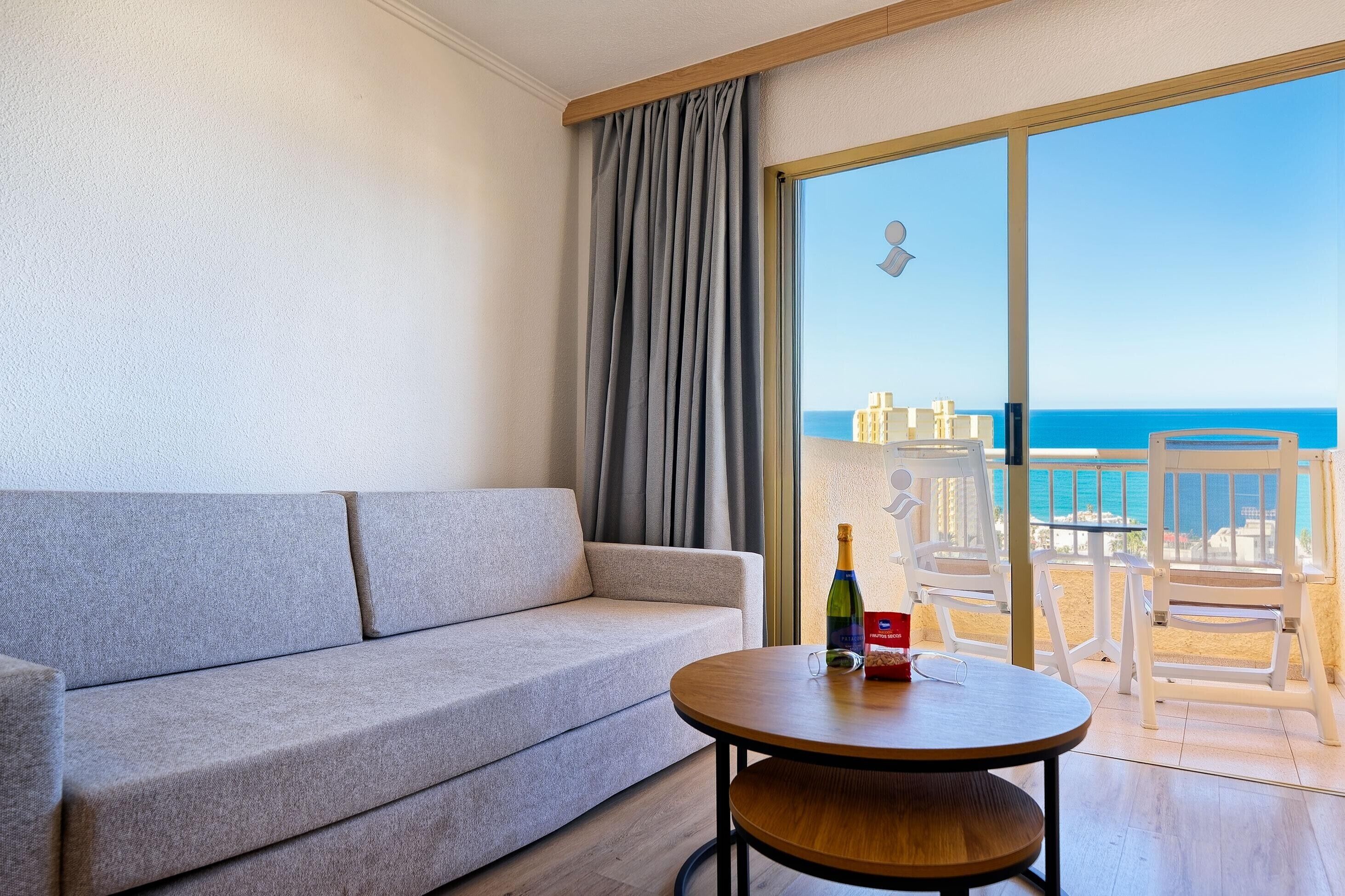 A room with a grey sofa, a round wooden table, and a view of the beach through a large window.