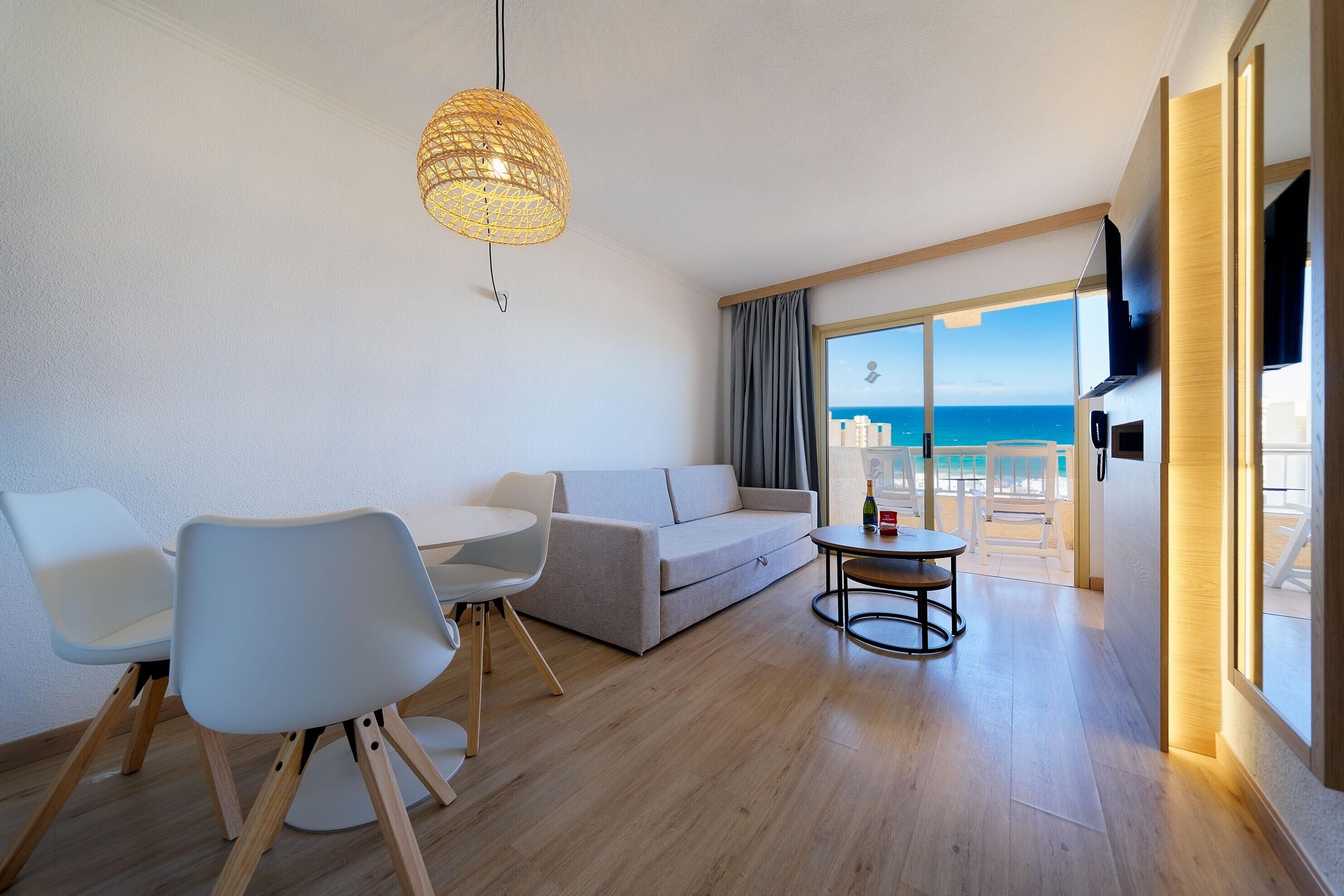 A modern living room with a sofa, chairs, a table, and a large window with a view of the sea.