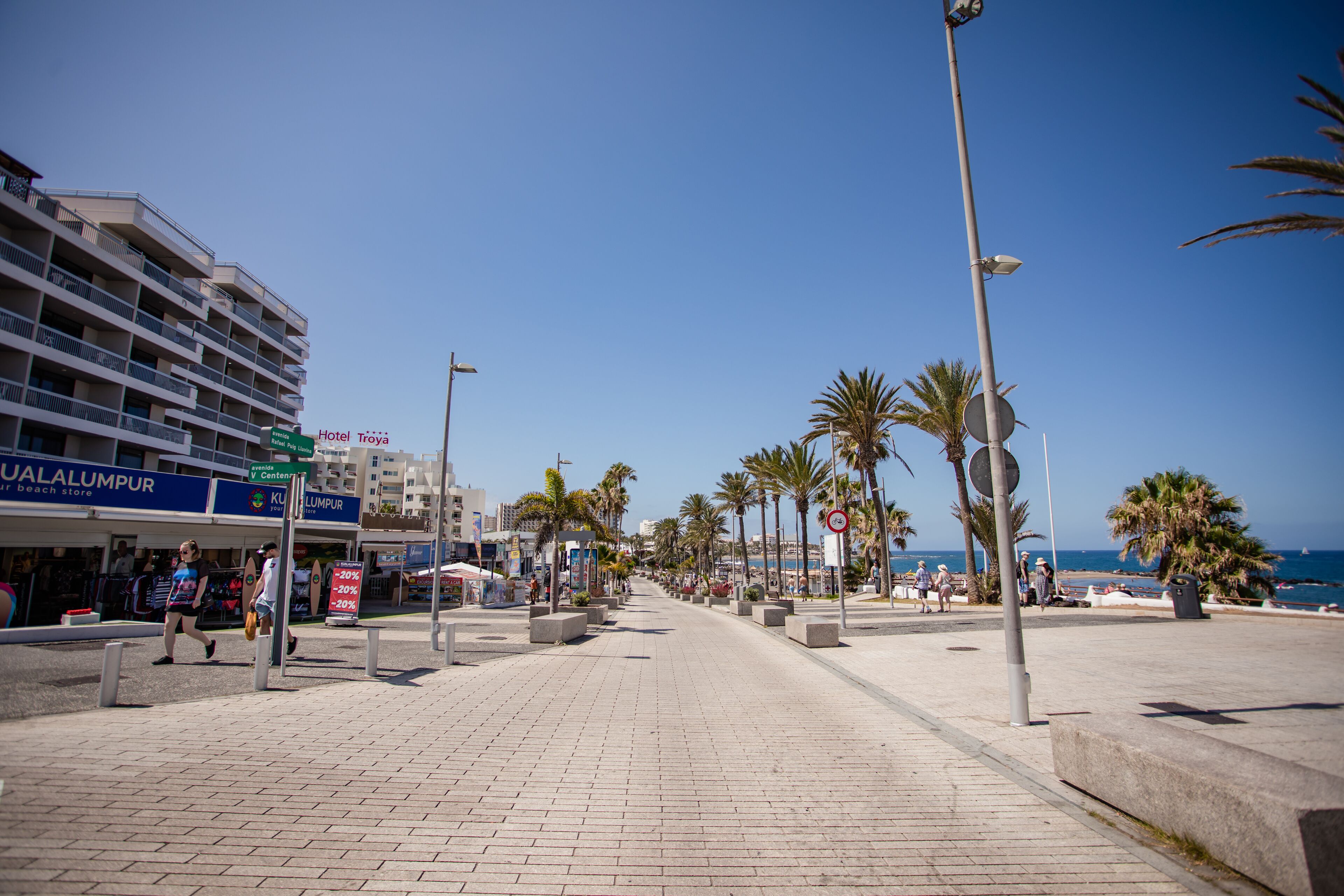 Un passeig marítim amb un hotel anomenat "Hotel Troya" i una botiga de platja.