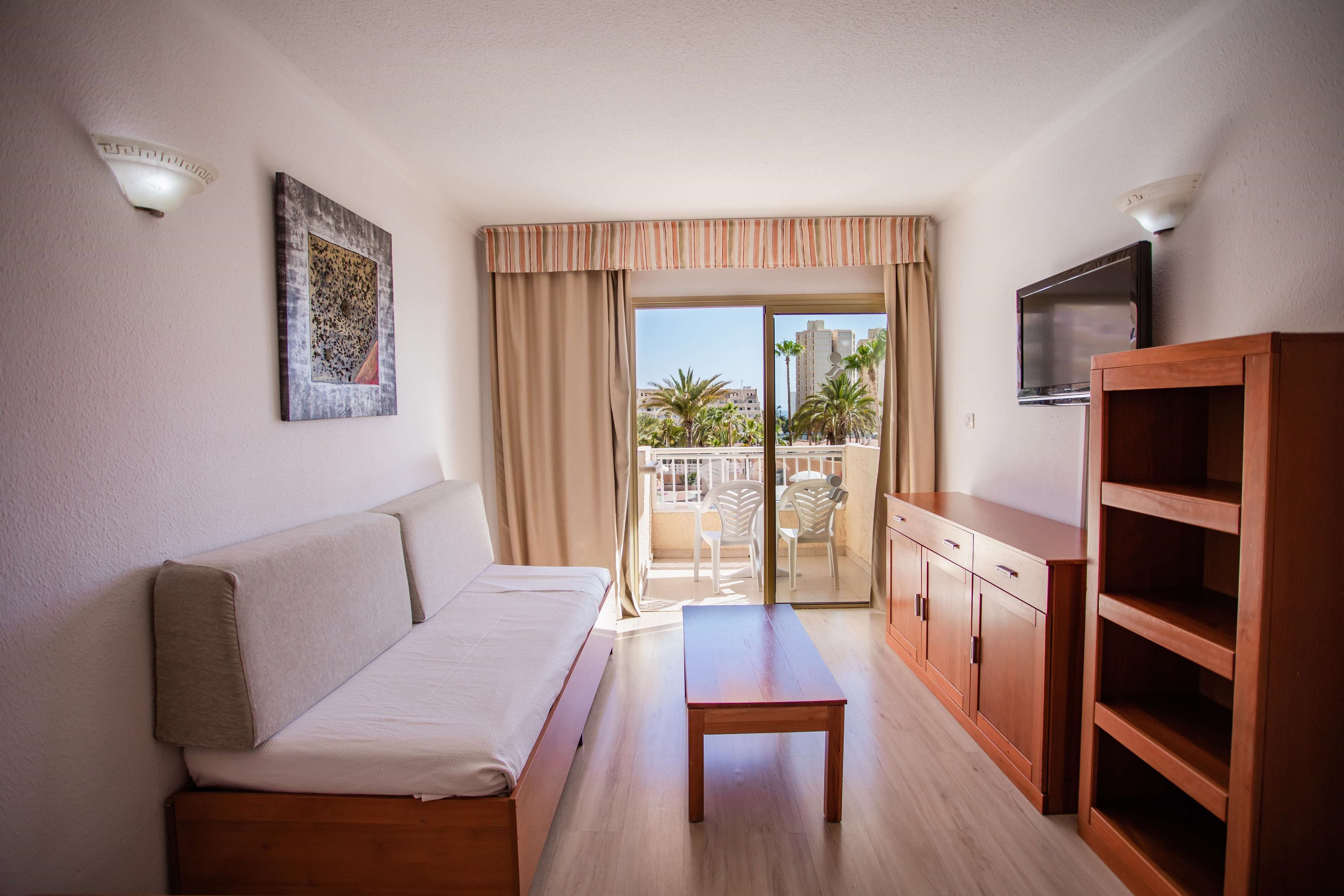 A hotel room with a sofa, a wooden table, a TV, and a balcony with a view of palm trees.