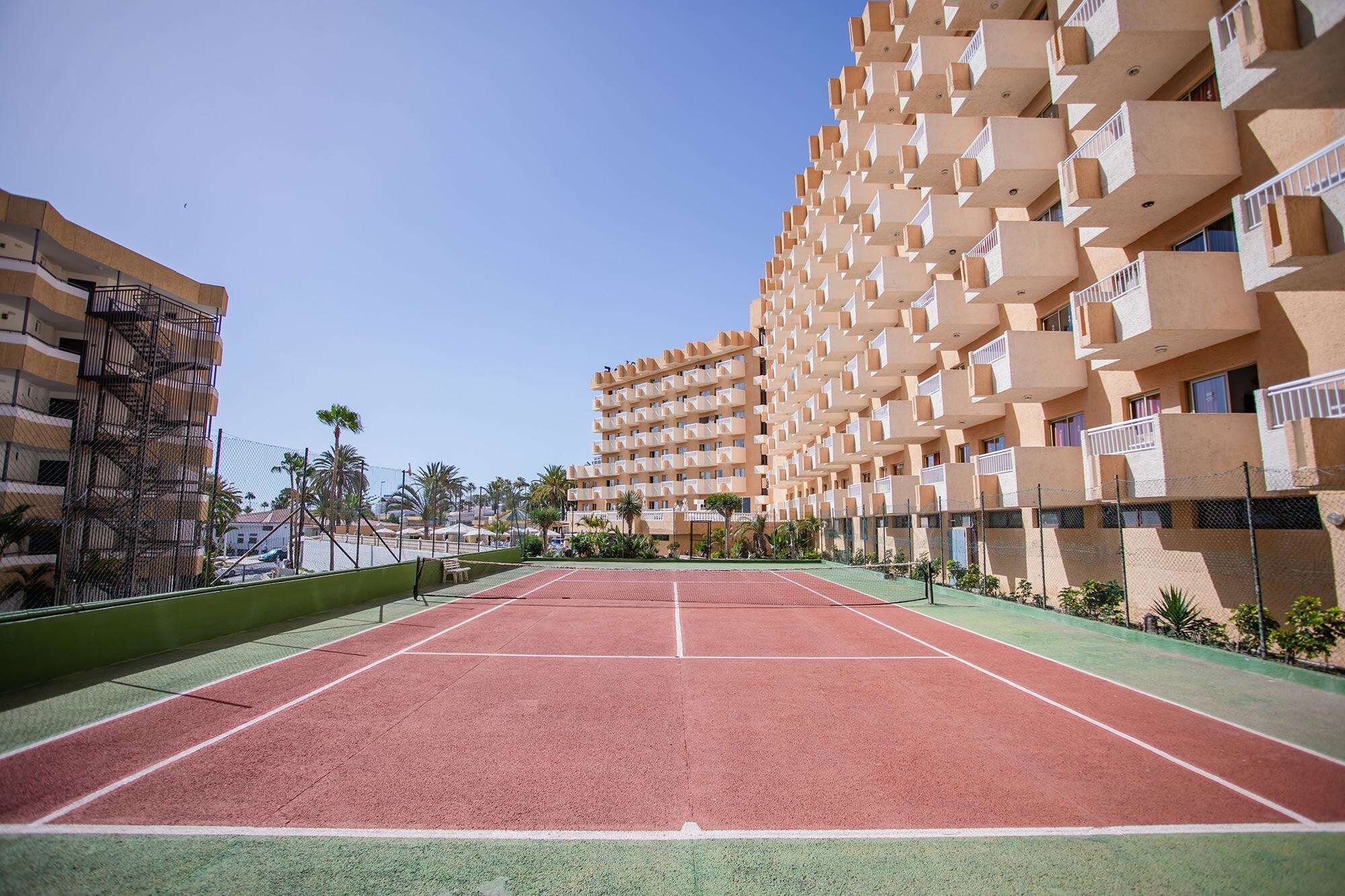 Una cancha de tenis con red, rodeada por una valla, y contigua a un edificio de varios pisos con balcones.