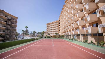 Una cancha de tenis con red, rodeada por una valla, y contigua a un edificio de varios pisos con balcones.