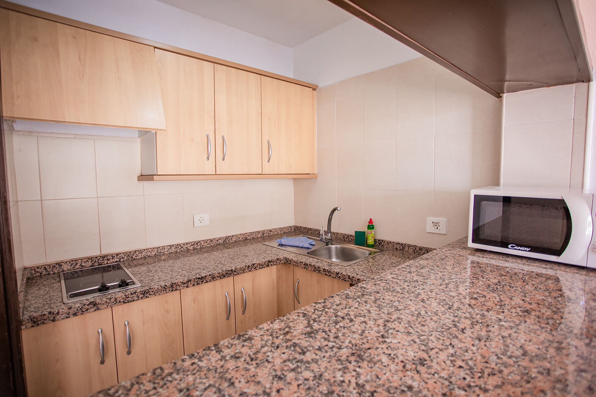A kitchen with granite countertops, wooden cabinets, a stainless steel sink, a microwave, and a dish soap bottle.