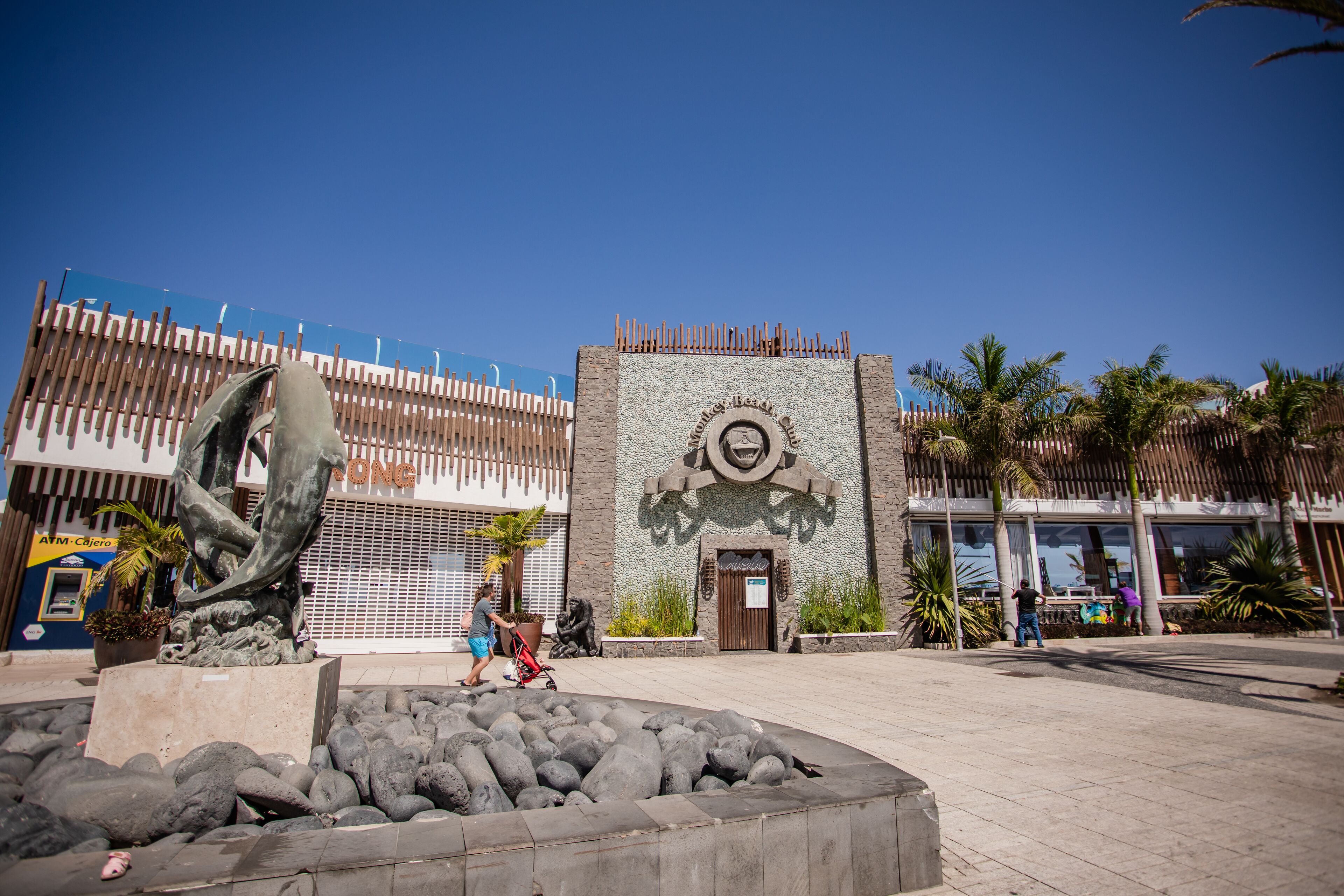 A plaza with a stone sculpture, a building with a mural, and palm trees.