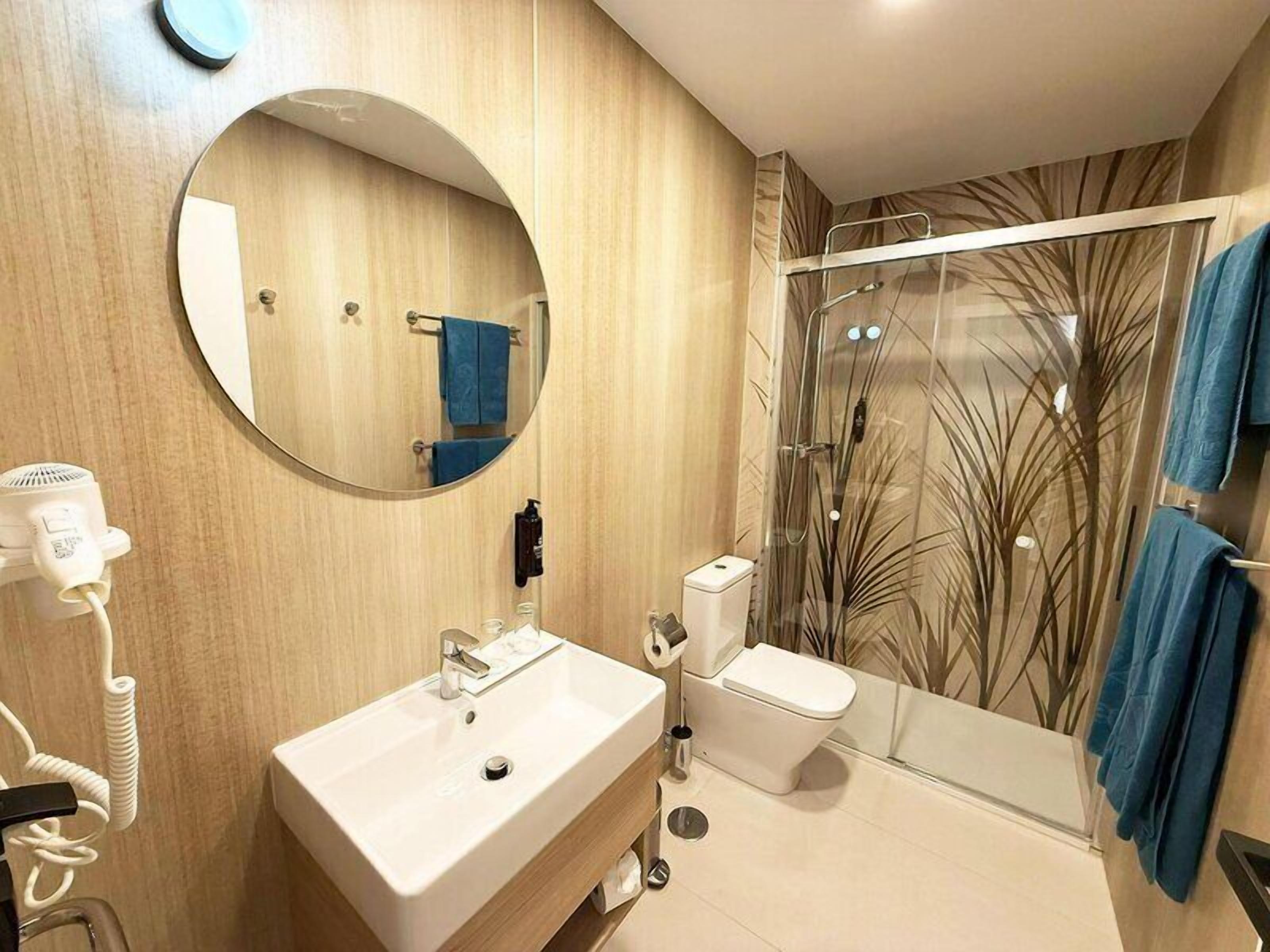 A modern bathroom with a round mirror, a white sink, a toilet, and a glass shower enclosure.