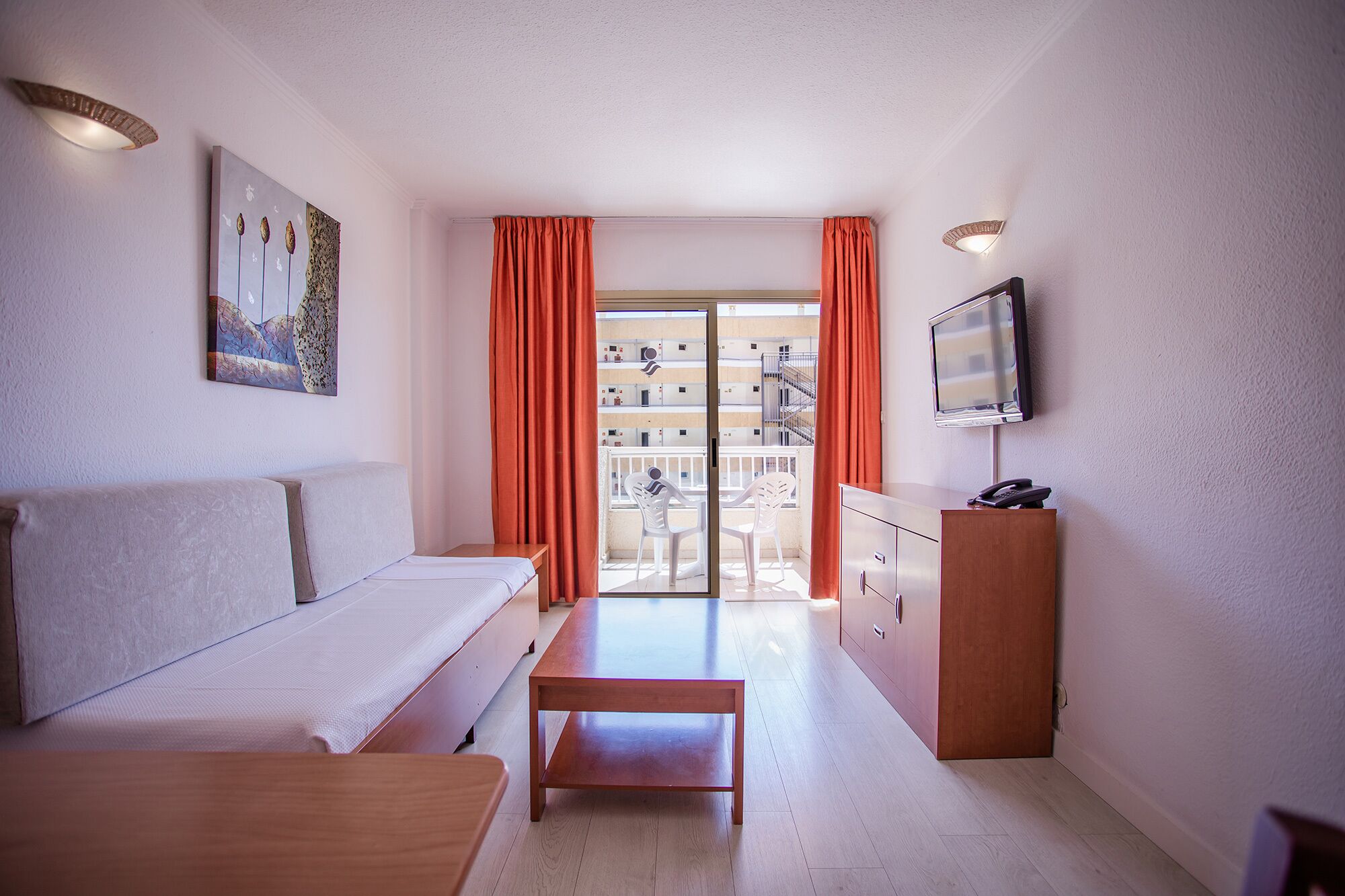 Camera d'albergo con un letto, una panca in legno, una scrivania con il telefono, una TV e un balcone con tende arancioni.