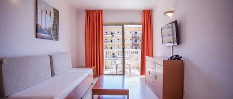 Camera d'albergo con un letto, una panca in legno, una scrivania con il telefono, una TV e un balcone con tende arancioni.