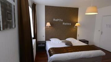 A hotel room with a bed, a headboard with 'Repos Reves' text, two bedside lamps, a nightstand, and a door.