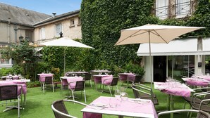 An outdoor dining area with tables and chairs, covered by umbrellas, set against a backdrop of a building with ivy.