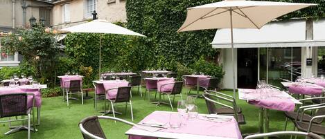 Un área de comedor al aire libre con mesas y sillas, protegida por sombrillas, frente a un edificio cubierto de hiedra.