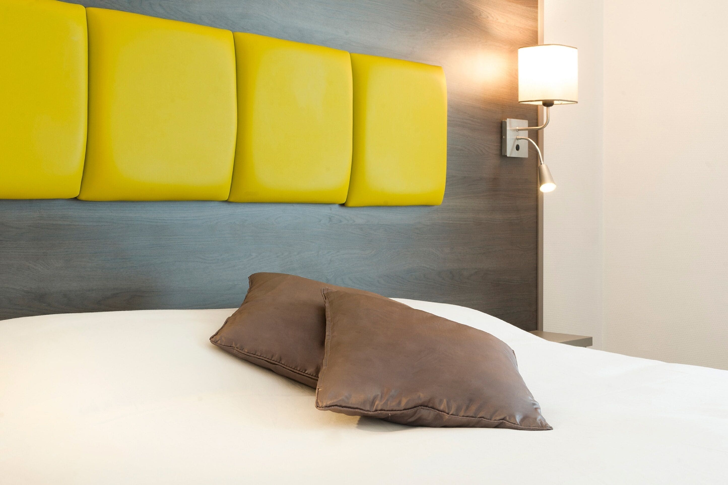 Una cama con dos almohadas, una pared con un panel amarillo y una lámpara de pared.