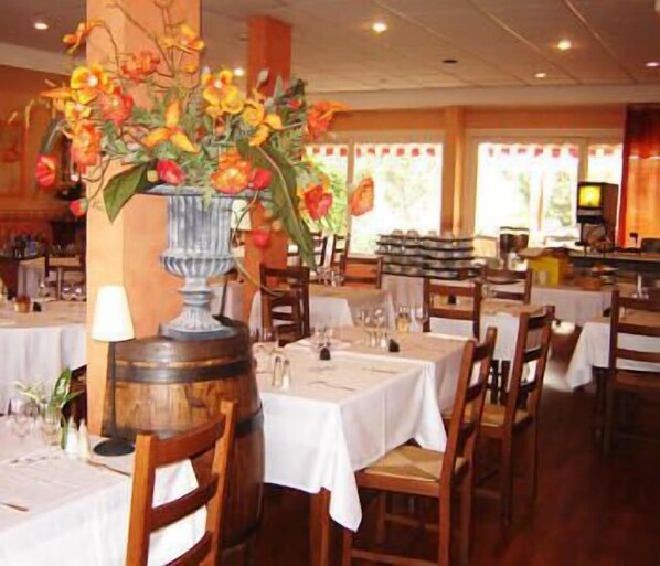 A restaurant interior with tables set for dining, a large vase of flowers, and a bar area in the background.