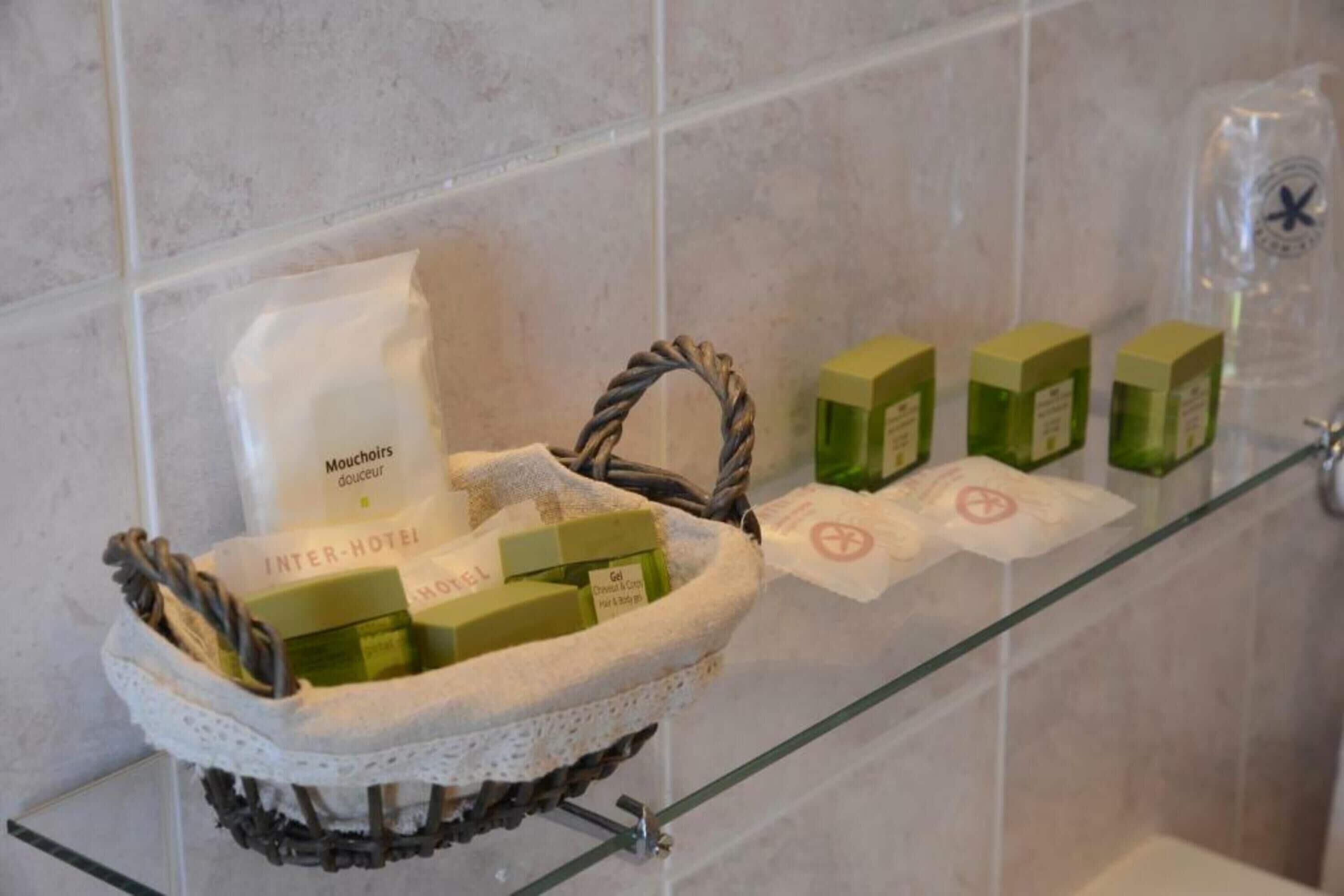 A bathroom with a glass shelf displaying toiletries and a basket with a bag labeled "INTER-HOTEL" and green soap bars.