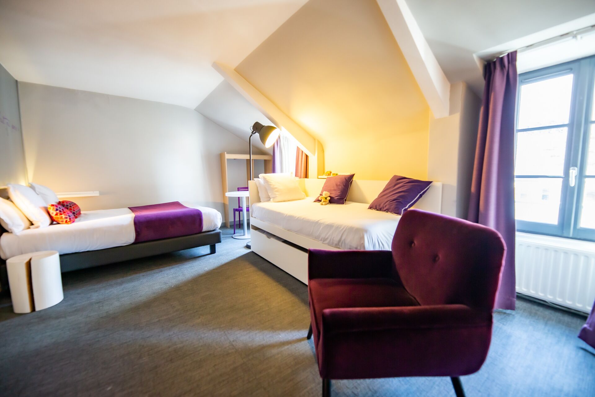 A modern hotel room with a loft bed, a daybed, a red armchair, and a small table with a lamp.