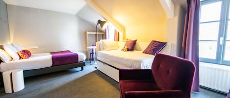 A modern hotel room with a loft bed, a daybed, a red armchair, and a small table with a lamp.