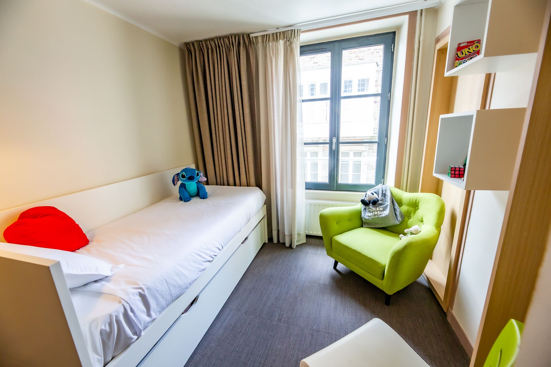 A hotel room with a single bed, a green armchair, a window with curtains, and a shelf with items.