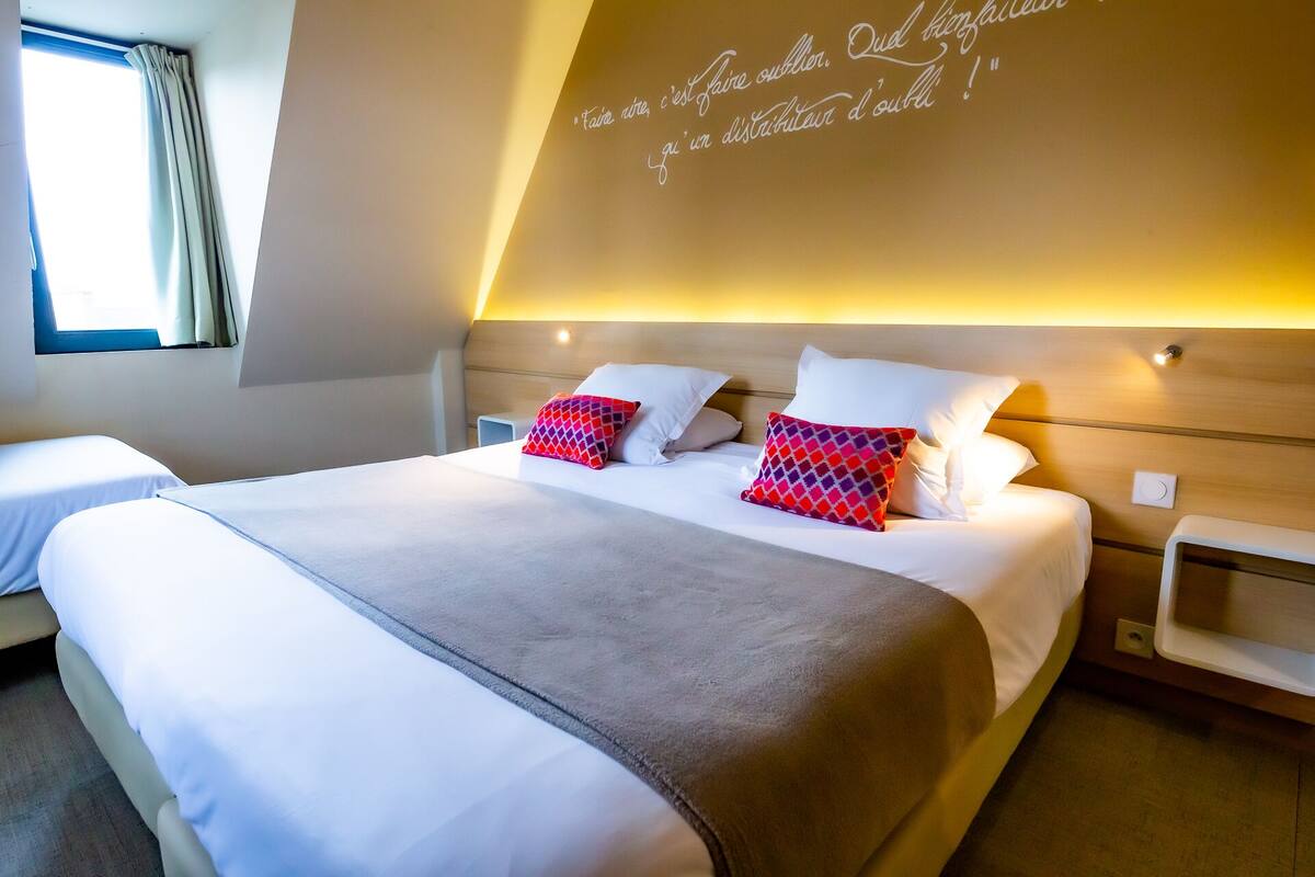 Habitación de hotel con una cama grande, dos almohadas, una mesita de noche y una pared con una cita.