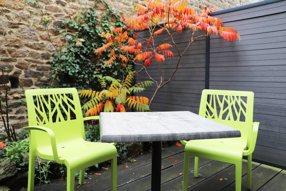 Una mesa y dos sillas con un diseño geométrico, ubicadas sobre una terraza de madera con un muro de piedra y follaje al fondo.