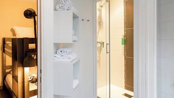 Un baño moderno con cabina de ducha de vidrio, un perchero blanco con toallas dobladas cuidadosamente y una cama con cabecera negra.
