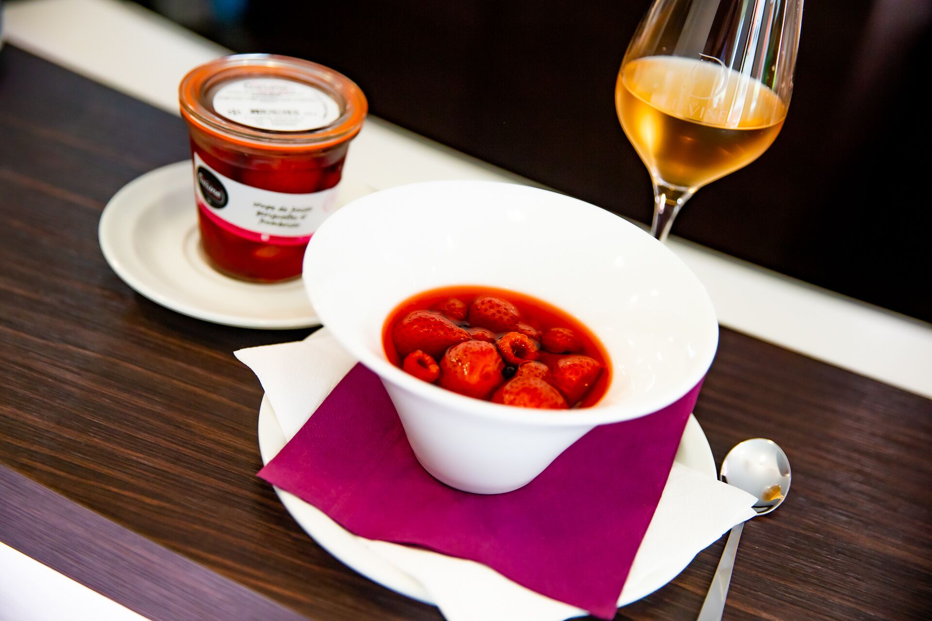 A bowl of strawberries with a spoon and a glass of wine on a table.