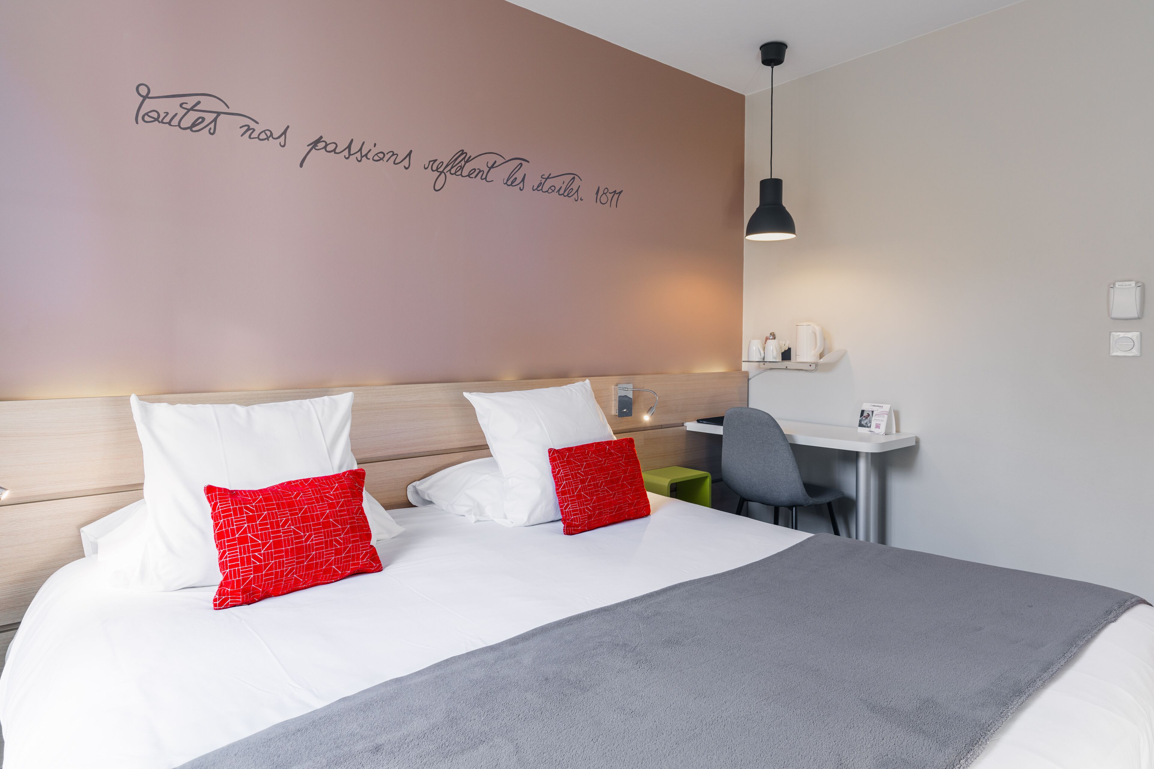 A neatly made bed with white and red pillows, a grey blanket, and a wall with a motivational quote.