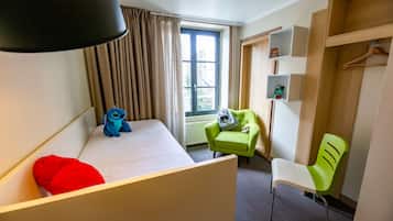 A hotel room with a bed, a green armchair, a white chair, a shelf with books, and a window with curtains.