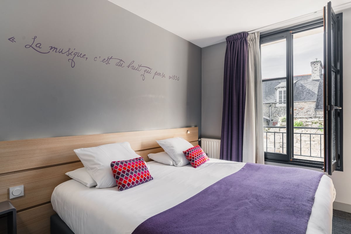 Habitación de hotel con una cama grande, cabecera, ventana con cortinas y una pared con una cita.