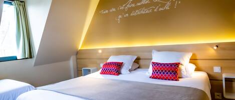 A hotel room with a large bed, two pillows, a bedside table, and a wall with a quote in French.