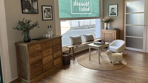 Een gezellige kamer met een houten dressoir, een witte fauteuil, een kleine tafel en een groot raam met uitzicht op de straat.