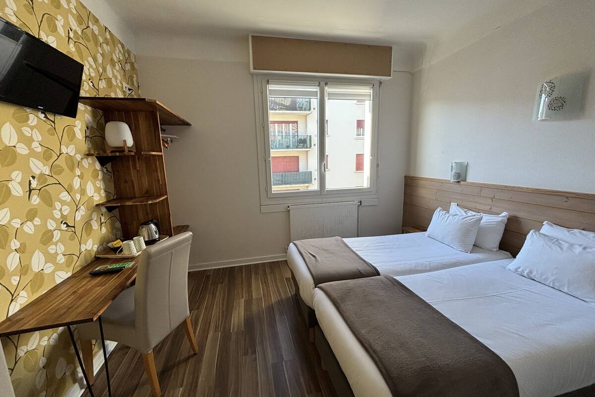 A hotel room with two beds, a desk, a television, and a window with a view of buildings.
