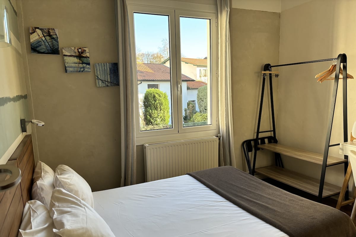 A bedroom with a bed, a window with curtains, a radiator, and a wooden shelf.