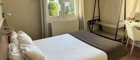 Una cama bien hecha con ropa de cama blanca y marrón, un escritorio pequeño con una silla y una ventana con vista a casas.