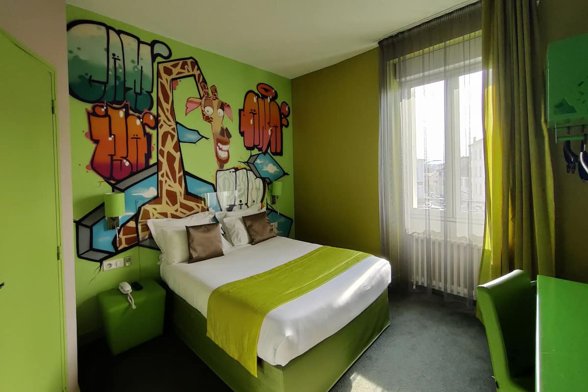Ein Hotelzimmer mit einem Bett, einer grünen Wand mit Graffiti und einem Fenster mit Vorhängen, sowie einem grünen Sessel.