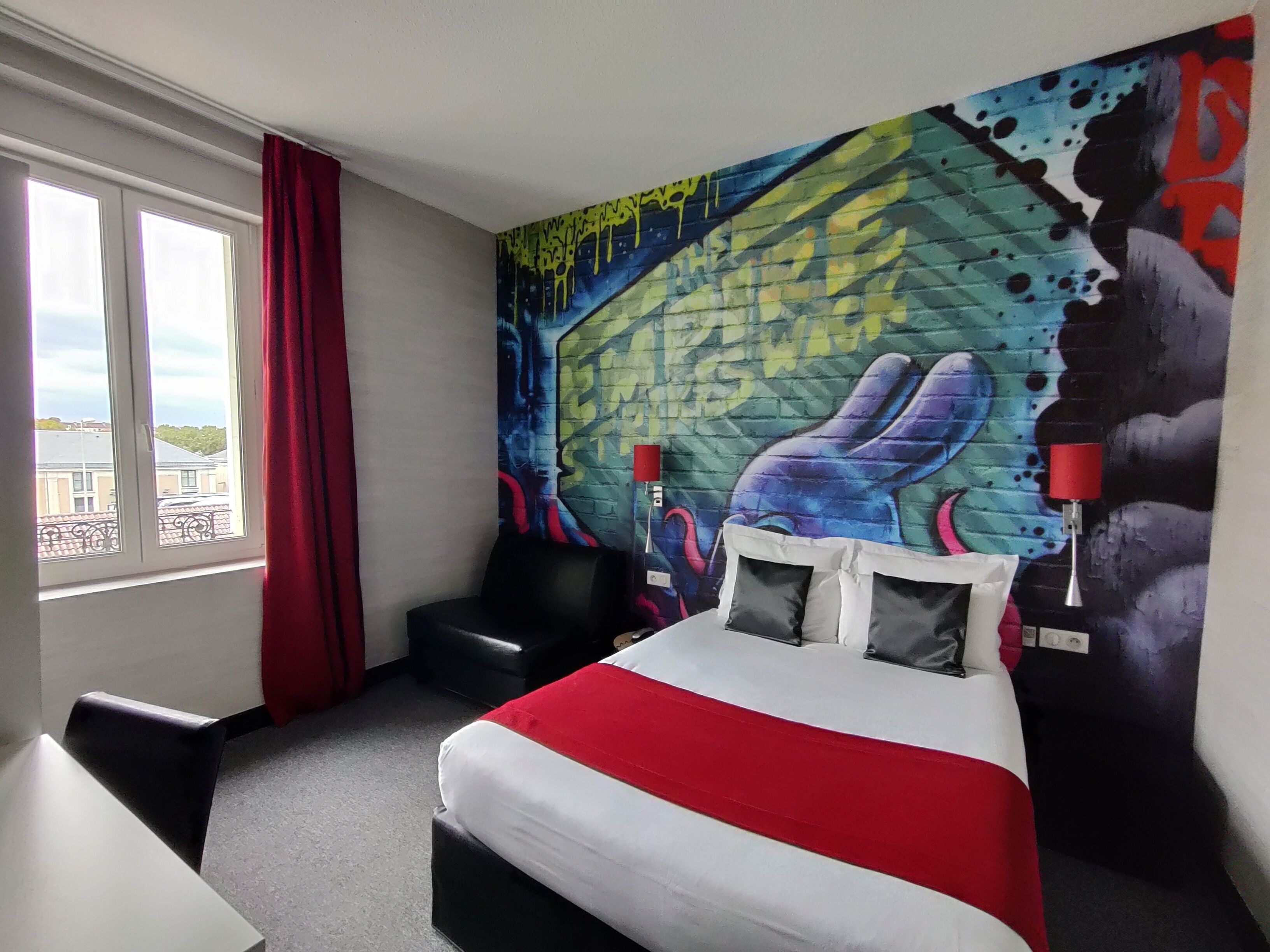 Ein Hotelzimmer mit einem großen Bett, einem Sessel, einem Fenster mit Vorhängen und einem farbenfrohen Wandgemälde.