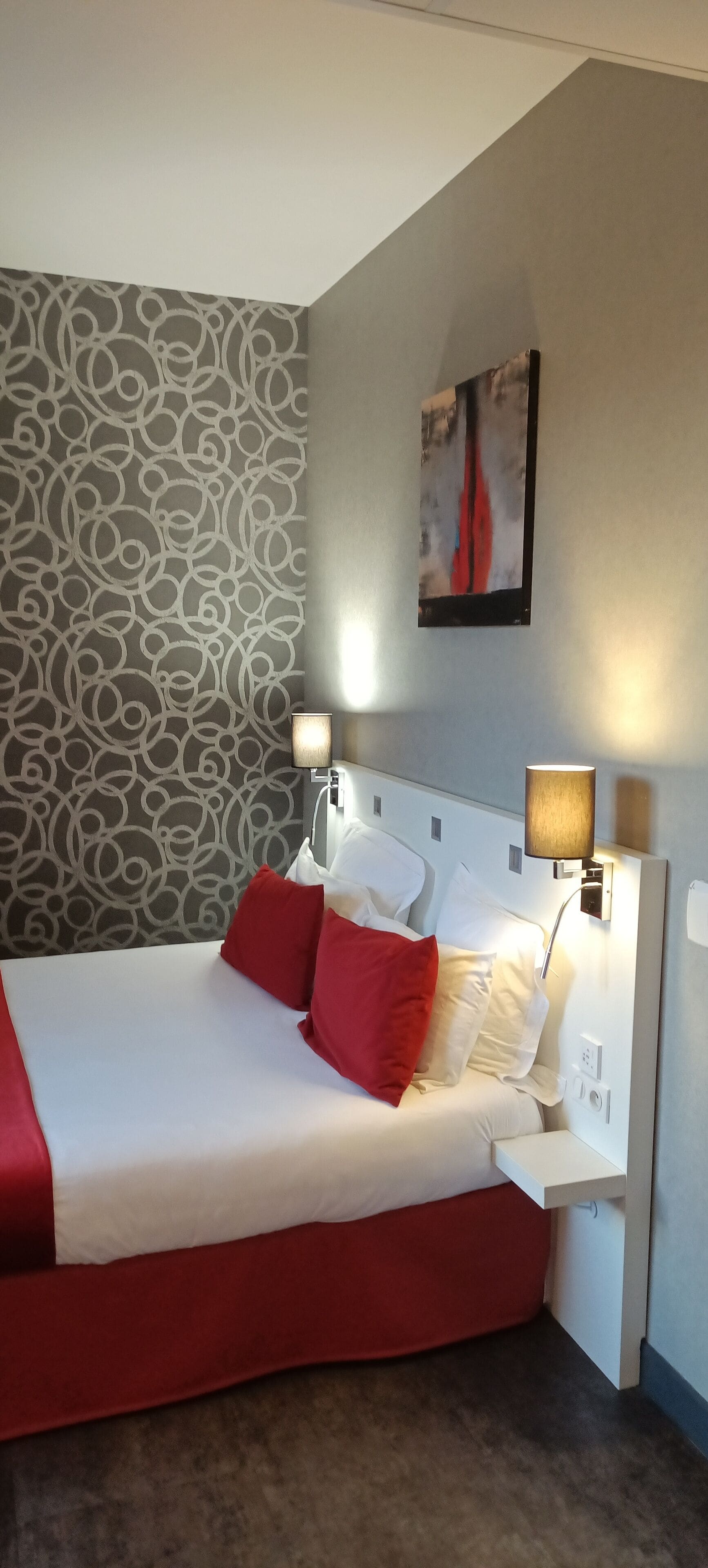 A neatly made bed with red and white bedding, a patterned wallpaper, and a painting on the wall.