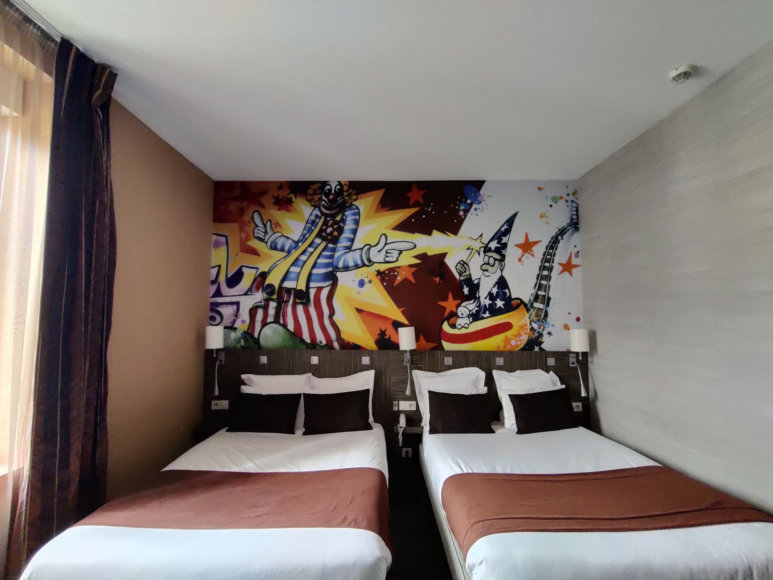 A hotel room with two beds, a colorful mural on the wall, and a window with curtains.