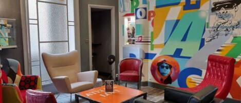 A room with a colorful mural, a beige armchair, a red armchair, a black sofa, and a small table with a sign.