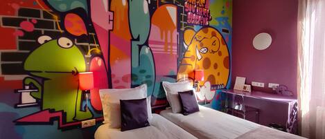 Habitación de hotel con una pared de grafitis de colores, una cama con almohadas, un escritorio con lámpara y una ventana con cortinas.