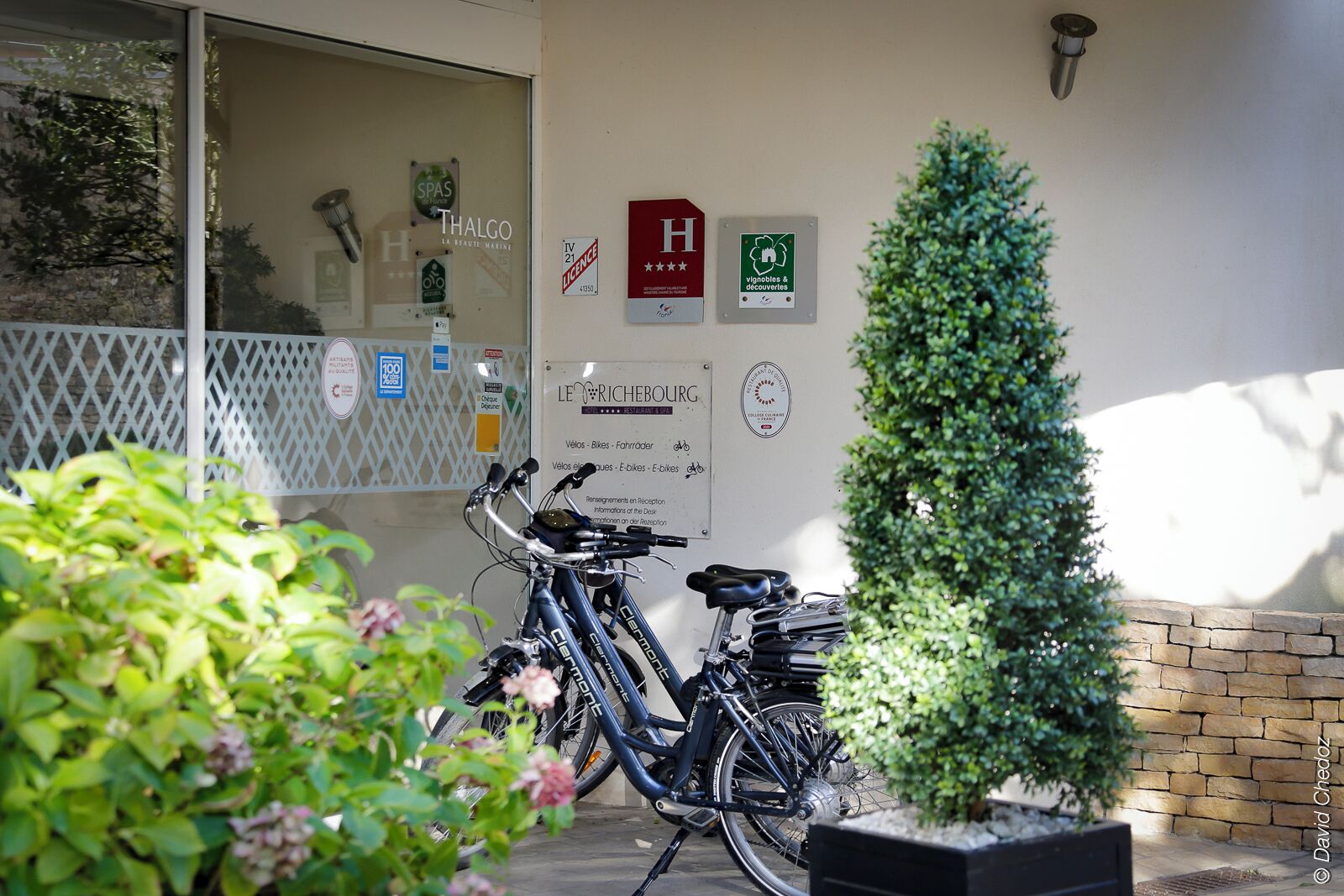 A building entrance with a sign for 'LE RICHEBOURG' and a 'Thalgo' sign, a small tree, and two bicycles parked outside.
