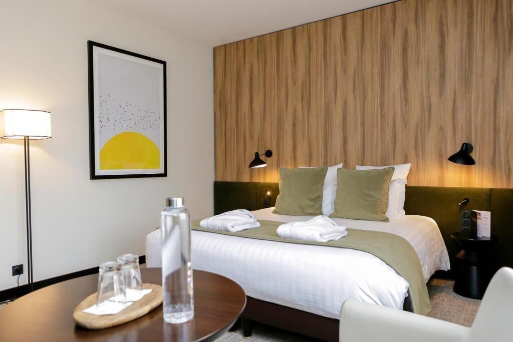A hotel room with a bed, bedside lamps, a framed picture, a table with glasses and a bottle, and a chair.