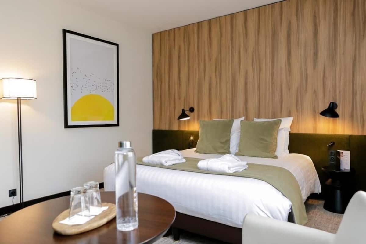 Ein Hotelzimmer mit einem Bett, Nachttischlampen, einem gerahmten Kunstwerk, einem Tisch mit Gläsern und einer Flasche sowie einem Nachttisch mit Lampe.