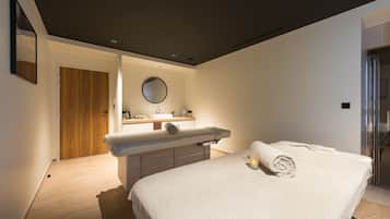 A modern bedroom with a wooden door, a round mirror, a bedside table, and a neatly made bed with a rolled towel.