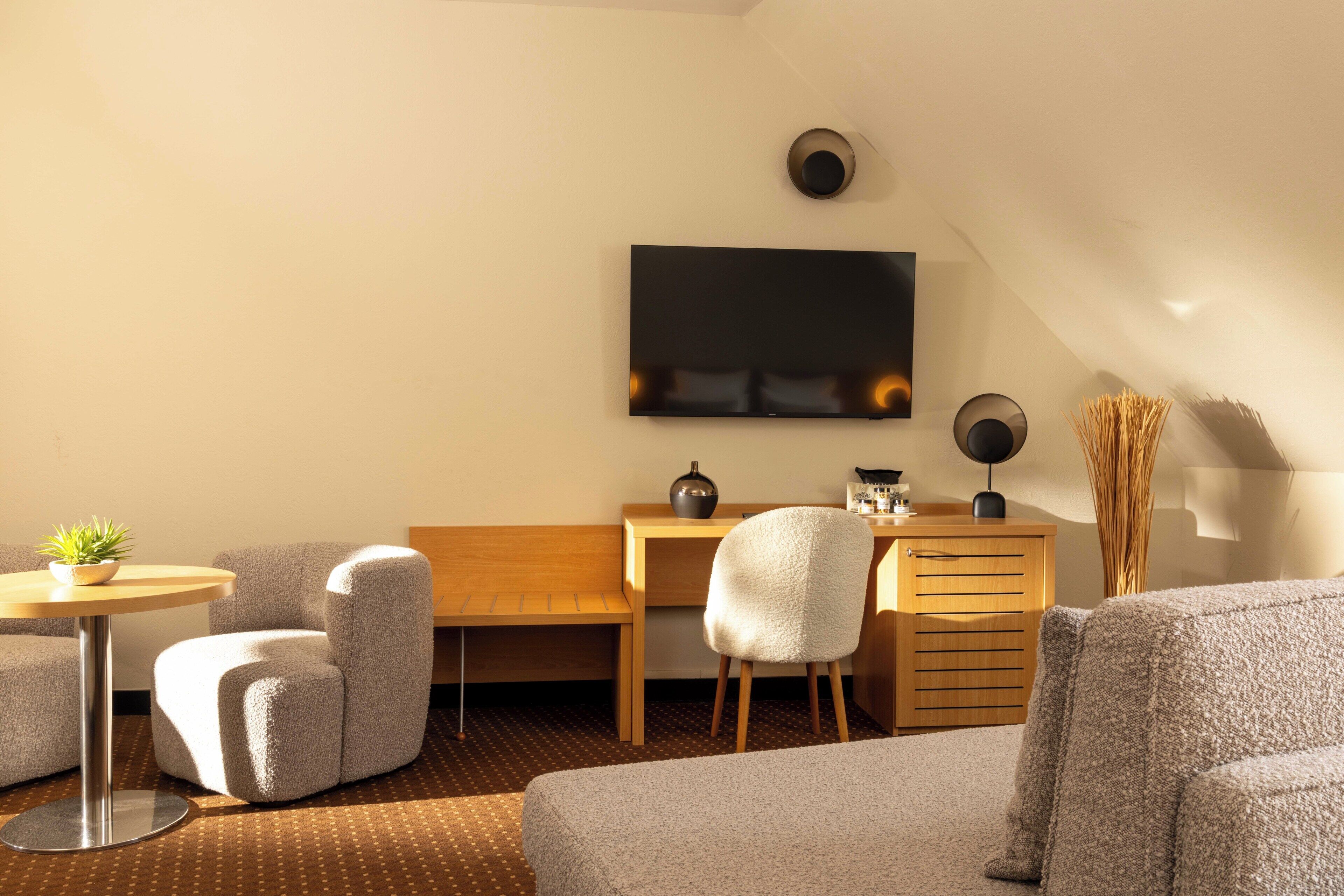 Una moderna camera d'hotel con divano, scrivania, sedia, un piccolo tavolo con una pianta e un televisore a schermo piatto montato a parete.