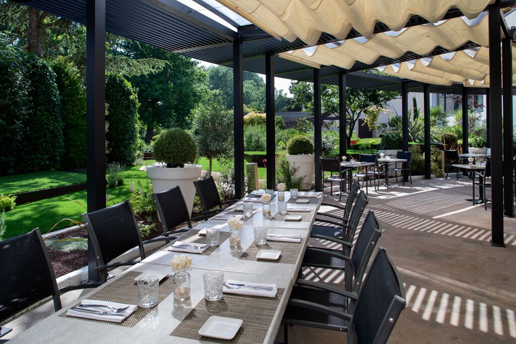 An outdoor dining area with tables set for six, surrounded by chairs, under a covered pergola with a view of a well-maintained garden.